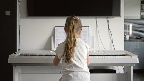 A 5-6 year old girl sits in front of a white digital piano and presses the keys. Back view.