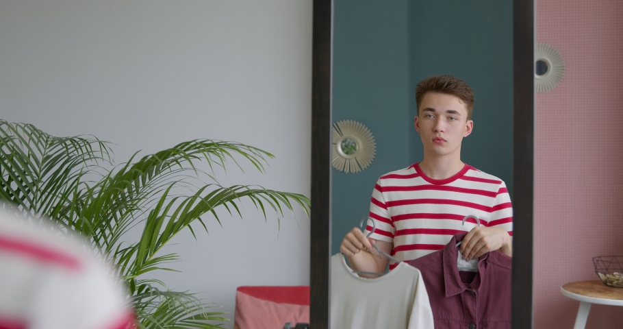 Young man is going to a meeting, chooses clothes in front of a mirror. Hard choice routine of what clothes to wear. Male hipster getting dressed looking reflection. Trying on clothes. Royalty-Free Stock Footage #1055943680