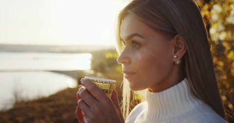 Young blonde happy woman is drinking coffee in cup while closing eyes with pleasure against of yellowed tree seashore in autumn orange sunset slow motion. Girl enjoys hot drink beautiful nature around