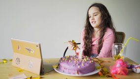 pretty little girl in pink plays with golden confetti having video call on tablet at wooden table with beautiful birthday cake in light room. Distant communication concept