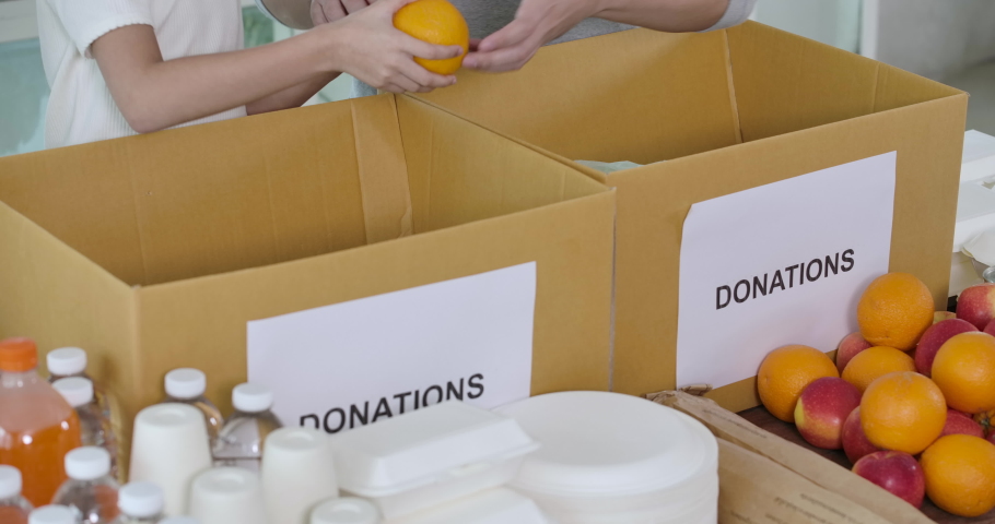 Close up shot of young food bank volunteers putting food products in donation box together as charity workers and members of the community work to the poor during the Coronavirus pandemic. Royalty-Free Stock Footage #1055951420