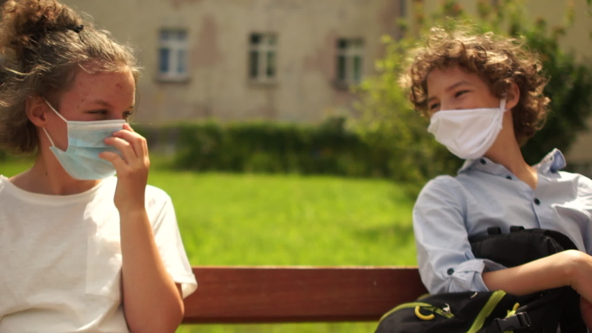 School-age children in medical masks sit on a bench and then say goodbye, touching each other with their elbows. Contactless farewell, back to school after quarantine coronavirus covid-19 Royalty-Free Stock Footage #1055951732