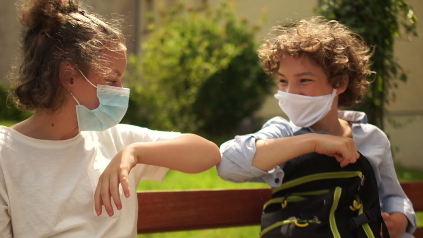 School-age children in medical masks sit on a bench and then say goodbye, touching each other with their elbows. Contactless farewell, back to school after quarantine coronavirus covid-19 | Shutterstock HD Video #1055951732