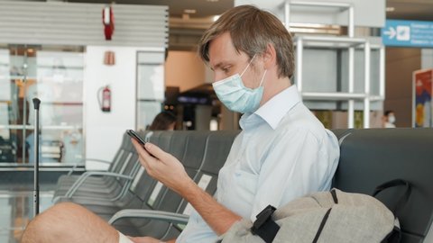 Business man Safe Travel freelancer in medical mask making a video call by mobile phone while waiting for his flight in airport terminal. Businessman doing a video conference call from his phone.