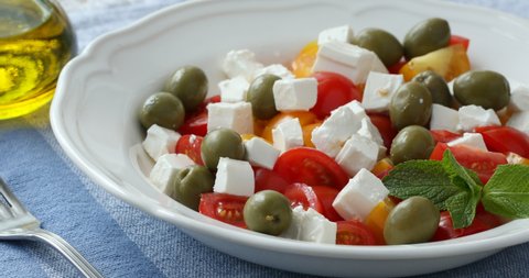 Pouring olive oil onto Greek salad with feta cheese, green olives, and cherry tomatoes, decorated with mint leaf