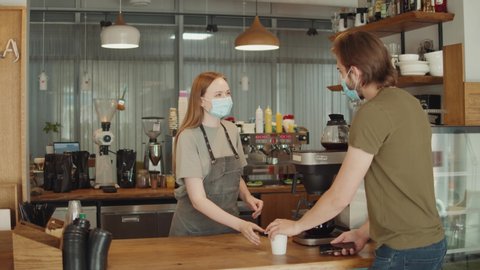 Barista with face mask in coffee shop, lockdown, quarantine, coronavirus is over, back to normal concept. Woman girl buy or purchase coffee contactless, banking terminal nfc payment by phone - Βίντεο στοκ