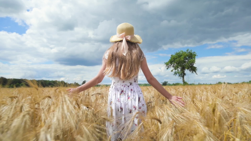 Beautiful happy Girl walking through golden Barley field and stroking ripe spikelets. Back view of cute Child in white dress and hat on cereal field. Slow motion.