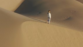 Traveler in bright casual sportswear is walking by the golden sand dunes with wavy texture. 4K aerial slow motion overhead video of a young woman at sunrise. Beautiful California nature landscape