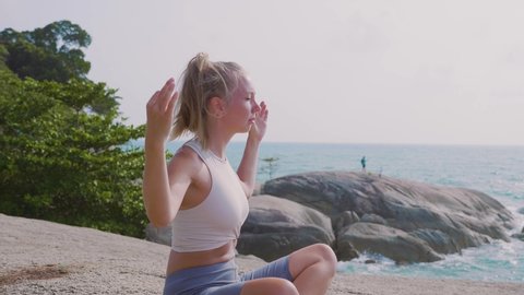 Pretty young slim woman meditating in lotus pose on the rocks by the tropical ocean beach, doing yoga on top of the mountain on a rock in nature. Healthcare, wellness, mindfulness concept.