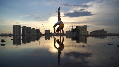 Flexible female gymnast doing acrobatic tricks with reflection in the water during dramatic sunset with cityscape background. Concept of fearless, courage and freedom 