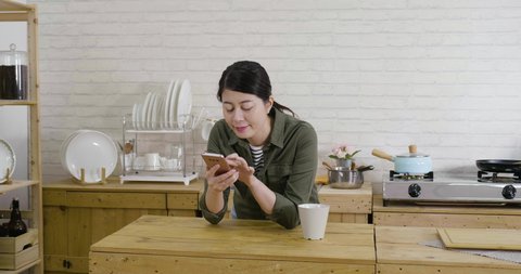 slow movement of young wife walk in cooking place at home. charming asian woman holding cellphone and cup of coffee leaning on wood table in kitchen. female enjoy morning tea and using mobile phone