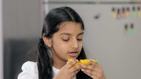 Closeup shot of a pretty young child adorably eating a slice of mango at home. A cute little girl enjoying tasty mango and showing a thumbs up while looking at the camera - juicy fruit concept