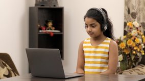 Young innocent kid busy doing a video call from her laptop - modern technology. Happy Indian girl waving a hand to her friend over a video call while sitting on a dining table in trendy summer wear