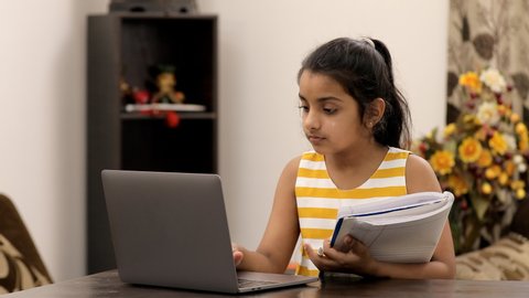 Medium shot of an adorable little kid studying online on a digital laptop at home. Beautiful Indian schoolgirl in casual wear, attending online classes via laptop - distance e-learning concept