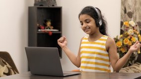 Medium shot - Cute little girl showing dance moves to her friends on a video call. Pretty Indian kid in headphones, happily dancing while doing a group video call during lockdown - distance communi...