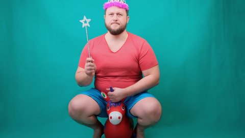 A younA young bearded freaky man in a pink T-shirt with a diadem on his head riding a unicorn with a magic wand in his hand. 