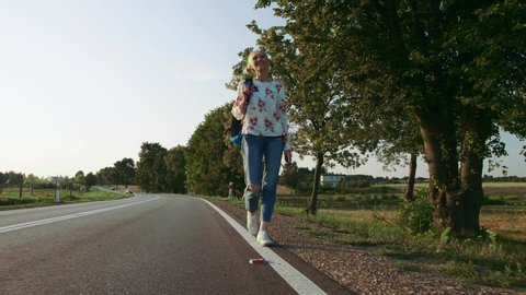 Woman with backpack walking on roadside. Cheerful young female with backpack walking on roadside while hitchhiking in Europe countryside during sunset.