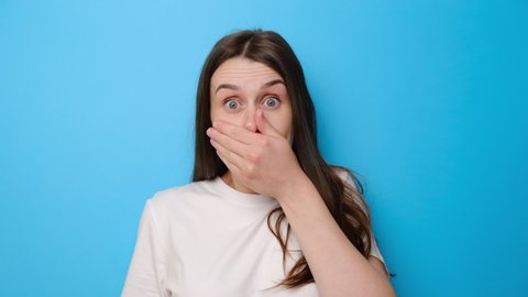 Shocked brunette girl isolated on blue studio background looking at camera with big eyes shocked expression, closing mouth zipping lips to keep terrible secret. People, emotions and unexpectedness