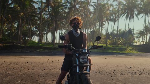 Portrait of a bold tattooed biker taking off bandana mask sitting on his motorbike. View on the beach against tropical palms. Free and self-reliant