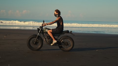 Concentrated man with face covered by a kerchief confidently driving his motorcycle by sandy ocean coast. Amazing wide waves on background changing by huge flat rocks lying near the water. Side view