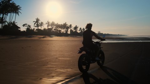 Scenic slow motion shot of man biker riding along the black sand beach of Bali in the warm light of evening sun. Feel free being away from hustle