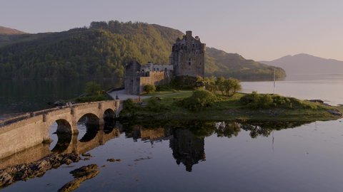 Aerial drone shot tracking around the stunning Eilean Donan Castle in Scotland at sunset, with the lake surrounding it and mountains in the background, and the reflection in the water.