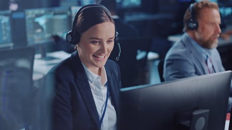 Happy Beautiful Technical Customer Support Specialist is Talking on a Headset while Working on a Computer in a Dark Monitoring and Control Room Filled with Colleagues and Display Screens.