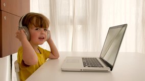Boy listens to music on headphones in front of laptop. Side view of child in wireless headset and casual clothes sitting at table in front of netbook with empty screen in apartment