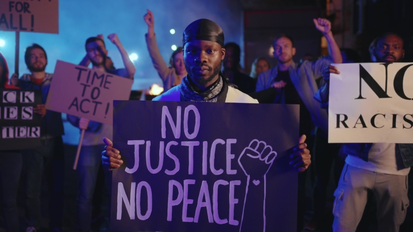 Portrait of black activist with No Justice No Peace placard among chanting crowd of protesters. Human rebellion against police violence in USA. Political rally. Revolution. Royalty-Free Stock Footage #1055988677