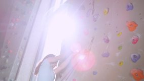 SLOW MOTION, CLOSE UP, LENS FLARE: Teenager skilfully climbs up a challenging route in a sunlit indoor rock-climbing training facility. Young Caucasian girl quickdraws her belay rope into a carabiner.