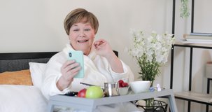Cheerful elderly lady smiling, waving and talking while using her smartphone for communication. Happy old woman in home robe having video conversation during breakfast in bed