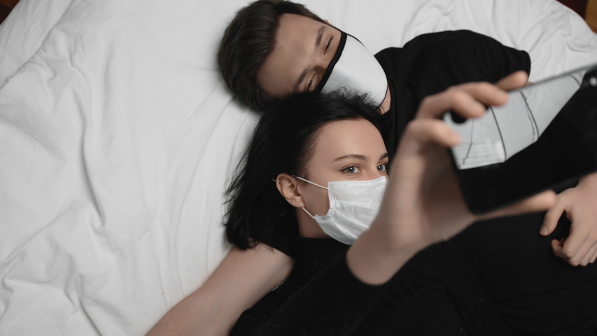 Cute couple, guy and girl use smartphone for video call or selfie. Attractive young woman and man in medical face masks lie on bed in quarantine or self isolation during coronavirus covid-19 pandemic Royalty-Free Stock Footage #1055991809