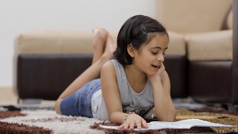 Young sincere schoolgirl reading her storybook at home - childhood education. Beautiful Indian kid in casual wear, reading her school book while lying on a carpet in the living room - kids lifestyle