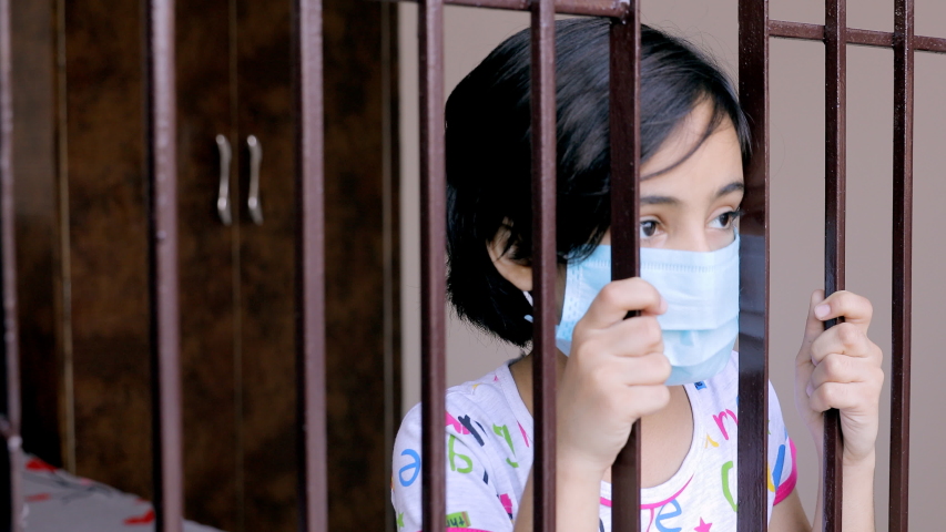Closeup of a young girl looking through the window while wearing a surgical mask. Portrait of a bored Indian kid in a medical face mask looks outside during the home quarantine - coronavirus pandemic Royalty-Free Stock Footage #1055994812