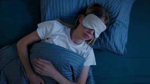 Young woman in bed in sleep mask, suffers from insomnia, tries to fall asleep, tosses, removes sleep mask from eyes and discontentedly look camera