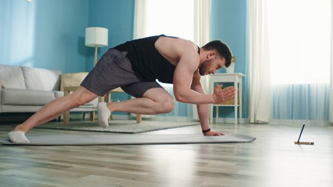 Young athletic man is training at home, using gadget, doing mountain climber exercise, working hard to maintain body shape in isolation, Zoom out, Slow motion.