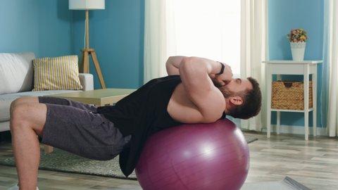 Young athletic man is workouting at home in the morning time, doing upper crunches, using stability ball, training hard to maintain his perfect body shape, Slow motion.