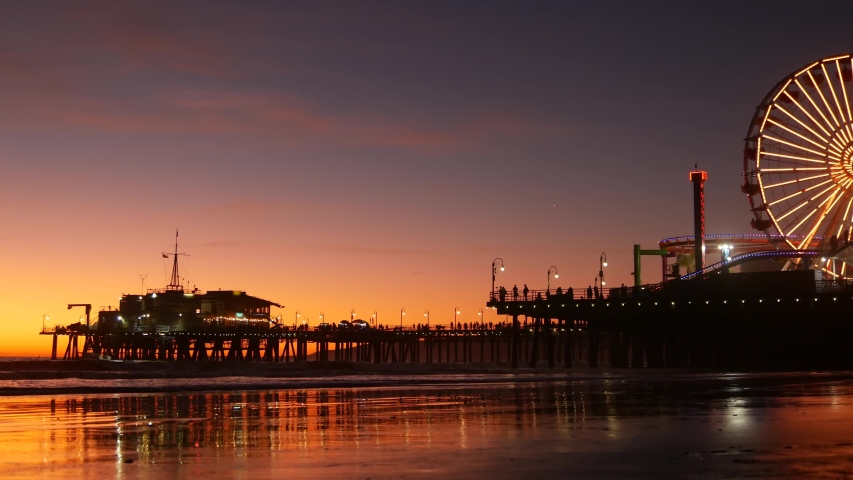 Twilight waves against classic illuminated ferris wheel, amusement park on pier in Santa Monica pacific ocean beach resort. Summertime iconic symbol of California glowing in dusk, Los Angeles, CA USA Royalty-Free Stock Footage #1055997899