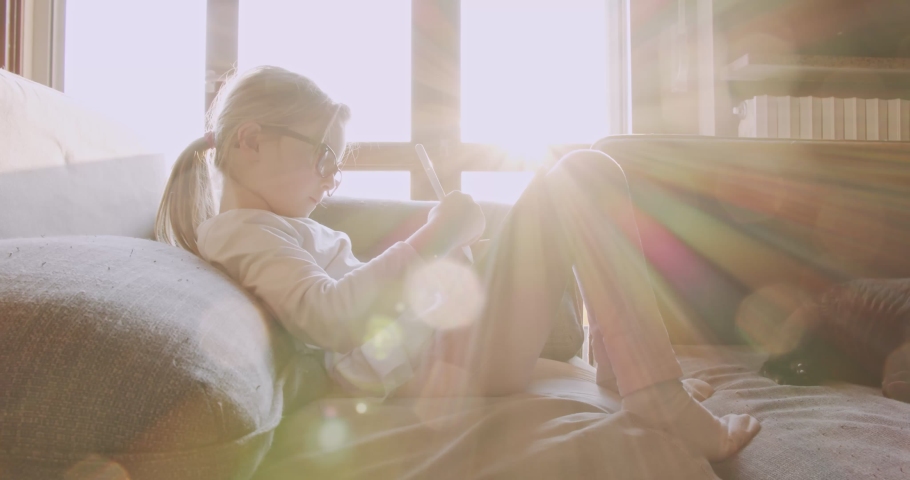 kid using tablet at home sitting on the couch. Little girl sitting on the sofa doing homework with tablet.Side view. Child playing with tablet in living room