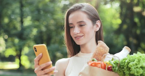 Millennial attractive woman hanging in social networks while standing in street and holding paper bag with groceries. Cheerful young woman scrolling news feed while using smartphone.