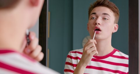 Young man queer holding lipstick or lip gloss in his hands and paints lips prepare getting ready in the morning. He looks at himself in the mirror. Get ready for a date.