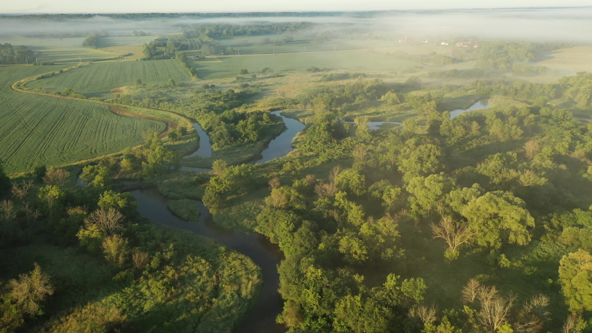 Aerial view of rural landscape with lush greenery, trees, river and fog mist above it. American countryside scenery in the early sunny morning, summertime. Midwestern nature. Camera moves upward