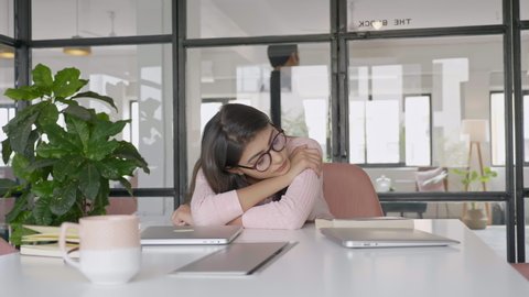 A beautiful and attractive office female girl employee with eyeglasses exhausted and tired sleeping or taking a nap on a desk during office or working hours in a modern corporate start up space. 