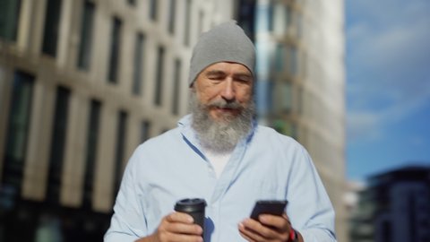 Panning medium shot of bearded senior man in hat walking down city street with takeaway coffee cup, text messaging on cell phone and thinking