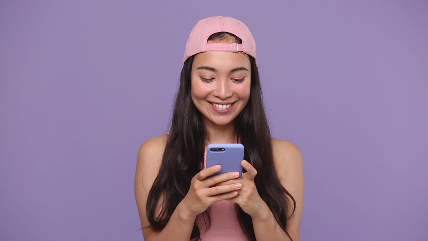 Young cute asian woman 20s in casual pink clothes cap isolated on pastel purple violet background studio. People lifestyle concept Look dreamful hold in hand using mobile cell phone send sms boyfriend