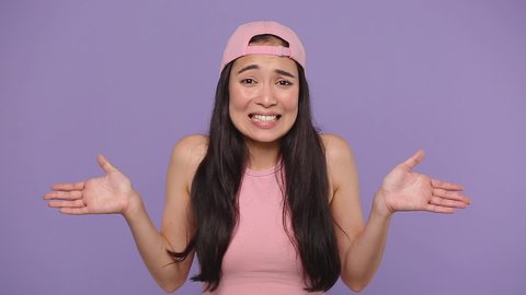 Fun confused shy shamed young asian woman girl 20s in pink clothes look camera bit lips spreading hands say oops i am sorry isolated on pastel purple violet background studio. People lifestyle concept