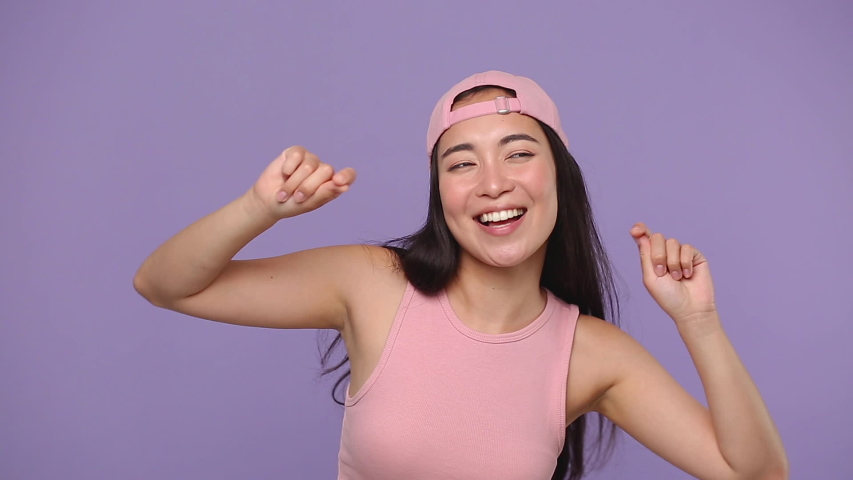 Young asian woman 20s in casual pink clothes cap posing isolated on pastel purple violet background studio. People lifestyle concept. Dancing fooling around having fun expressive gesticulating hands | Shutterstock HD Video #1056010826