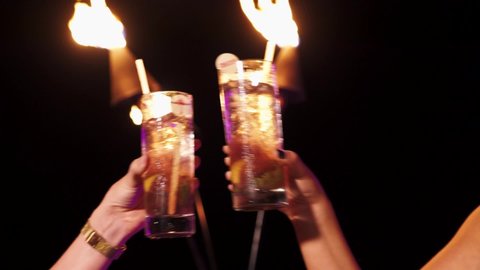 Close up of two drinks clinking glasses, cheers in front of lit tiki torches. 