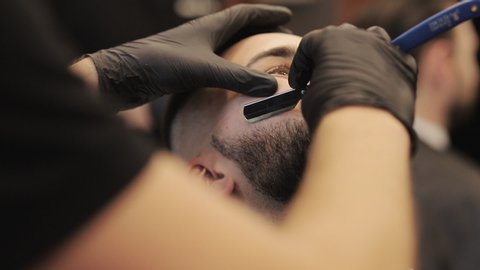 Professional barber shaves customer beard with straight razor. Beard cut with old-fashioned blade at barbershop. Handsome man getting his beard shaved in hair studio.