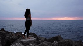 Barefoot girl practicing yoga in front of the mediterranean sea while the sun is almost on the horizon and shining with an orange light while standing on a rock